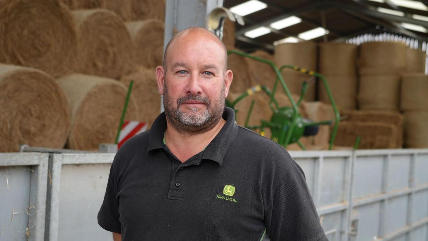 A portrait photo of sheep farmer Nick Whitehead inside a barn with hay bales and machinery in the background