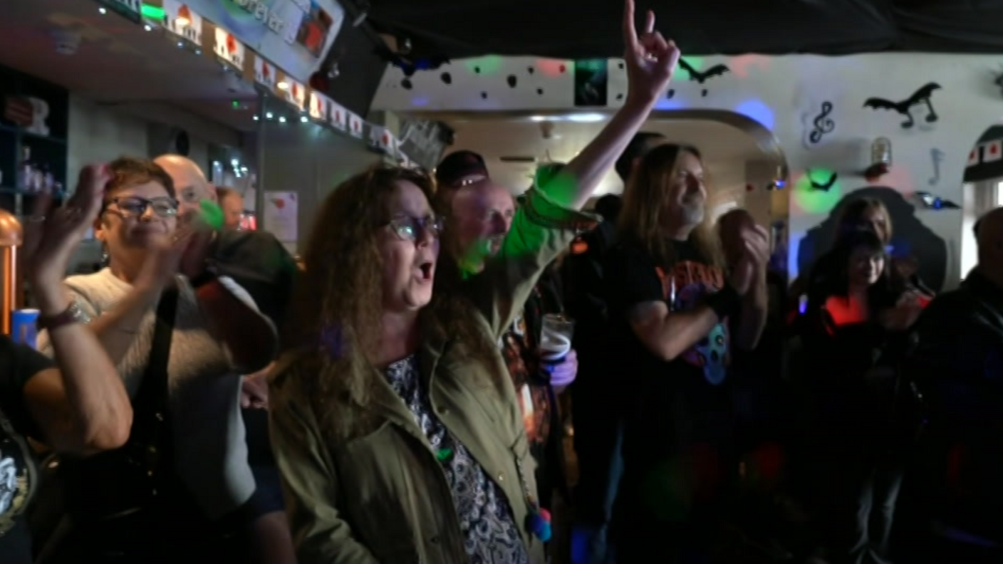 People are stood in front of a pub bar, watching a performance on stage. One woman has left arm raised in the air, her fingers pointing upwards