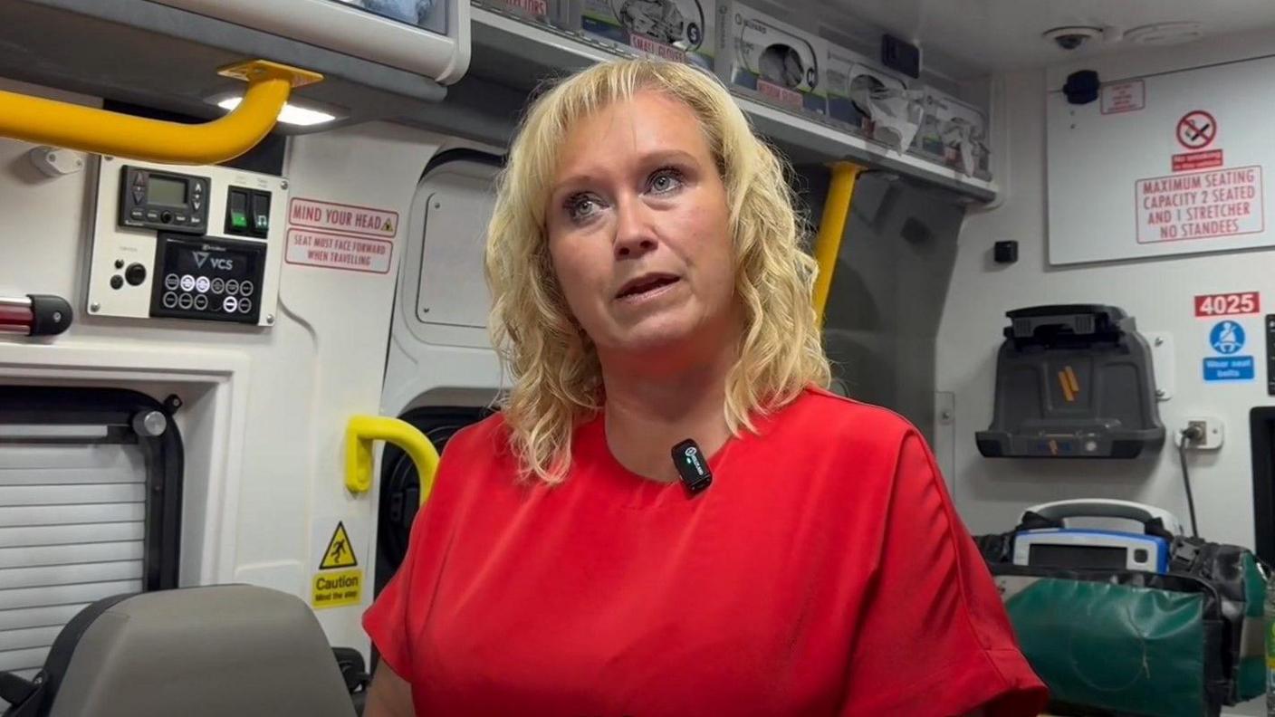 A woman with blonde shoulder-length hair, wearing a red top. She is standing in an ambulance and speaking in a promotional video for the scheme.  