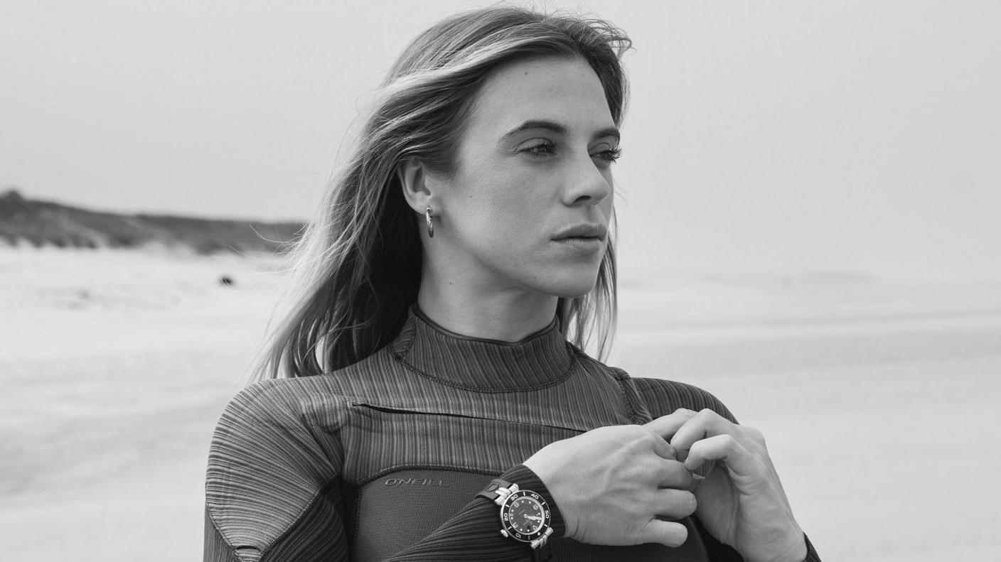 A black and white image of Laura Crane posing at the beach, with waves in the background. She is wearing a black O'Neill wetsuit and has long blonde hair and a black watch.