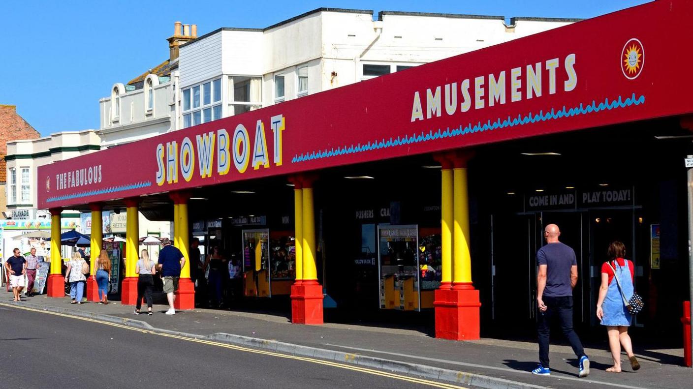 People walk past a seafront arcade with red sign and yellow pillars.