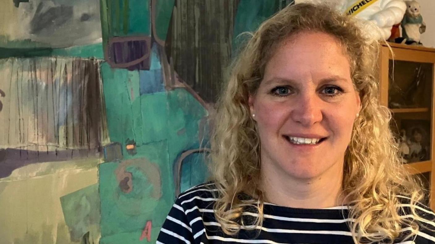 Camilla Notley has curly blonde hair and is smiling. She is standing in front of one of Malcolm Race's abstract paintings. Teal and turquoise shapes with grey lines. 