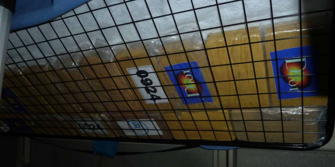 A number of bags, containing drugs, under the metal frame of a mattress in a lorry. They have numbers on the and the word Dior. The bags are yellow in colour. 