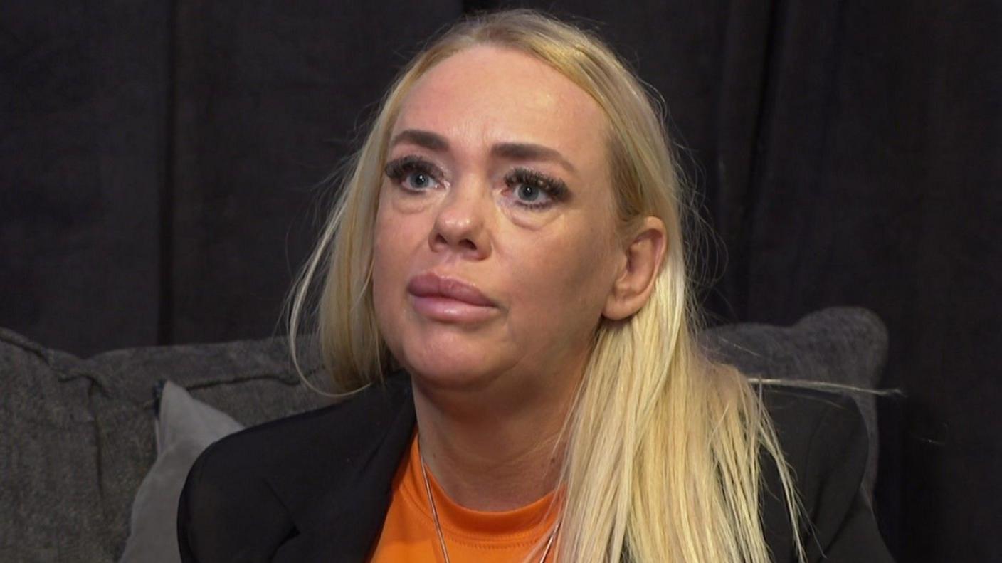 Heather Lane has long blonde hair. She is wearing an orange t-short and a black blazer.