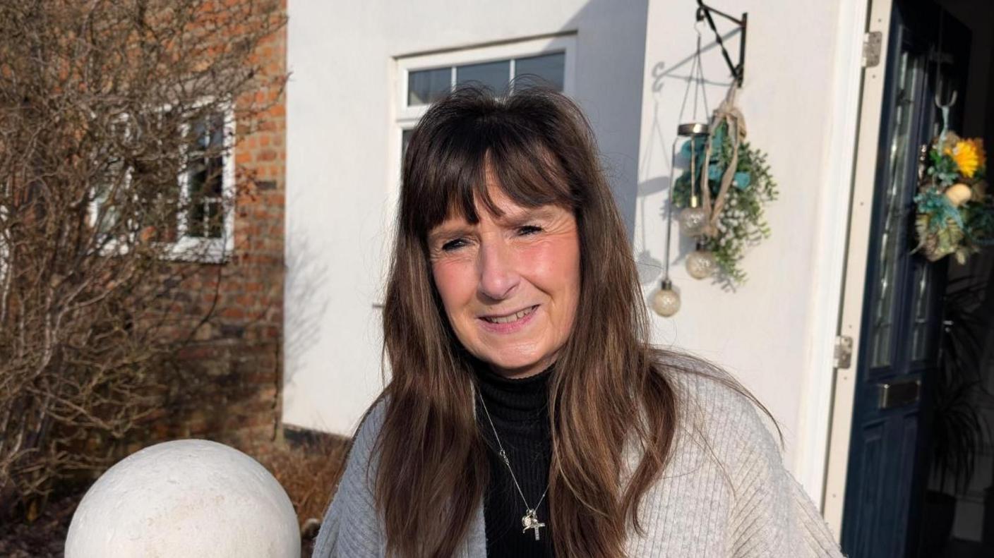 Head and shoulders shot of a woman standing in front of her house. She has long brown hair and wears a black top and grey cardigan