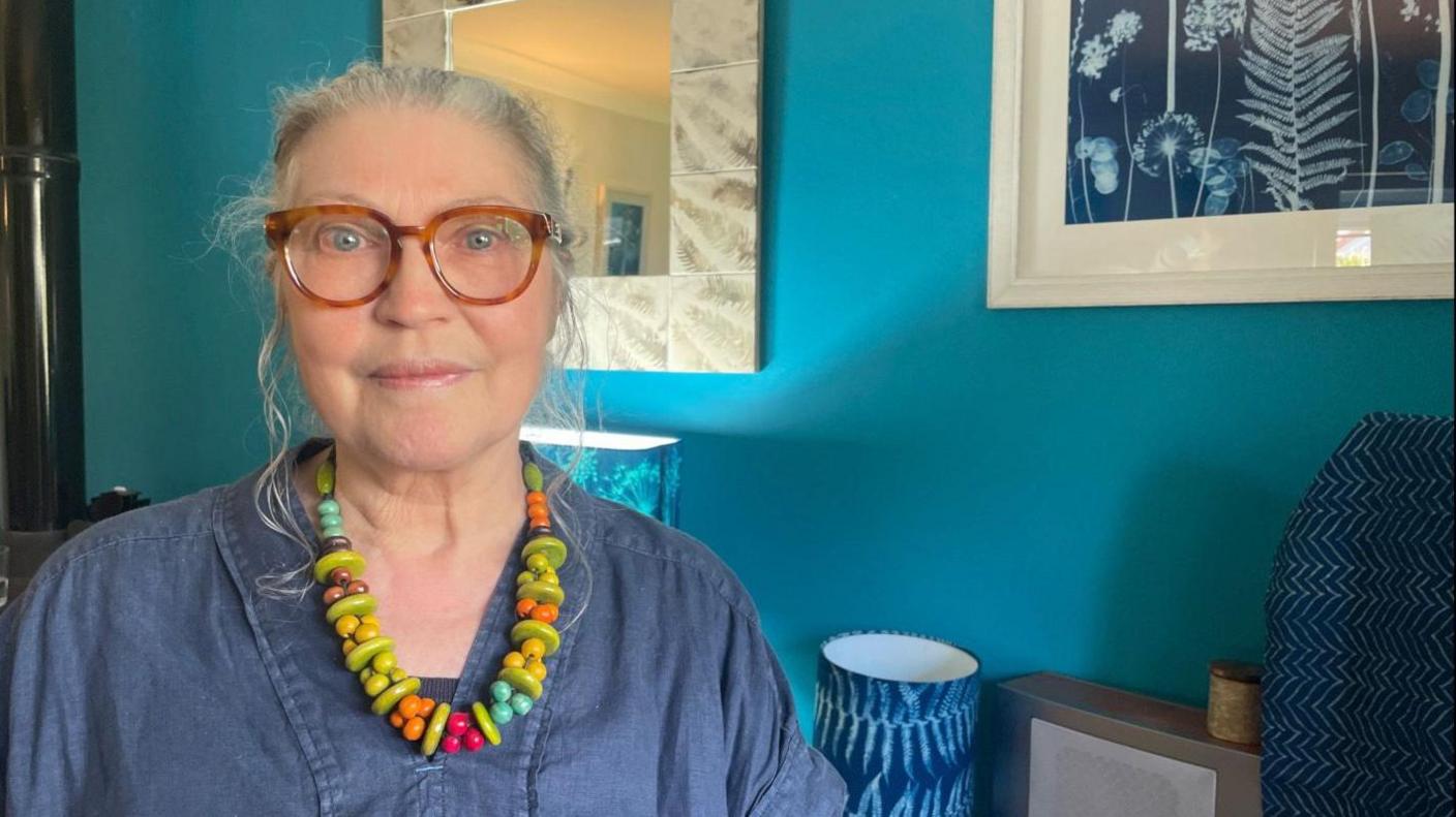 An artist with a colourful beaded necklace and glasses sits in front of a turquoise wall which has a cyanotype print on it by the artist Bridget Arnold.