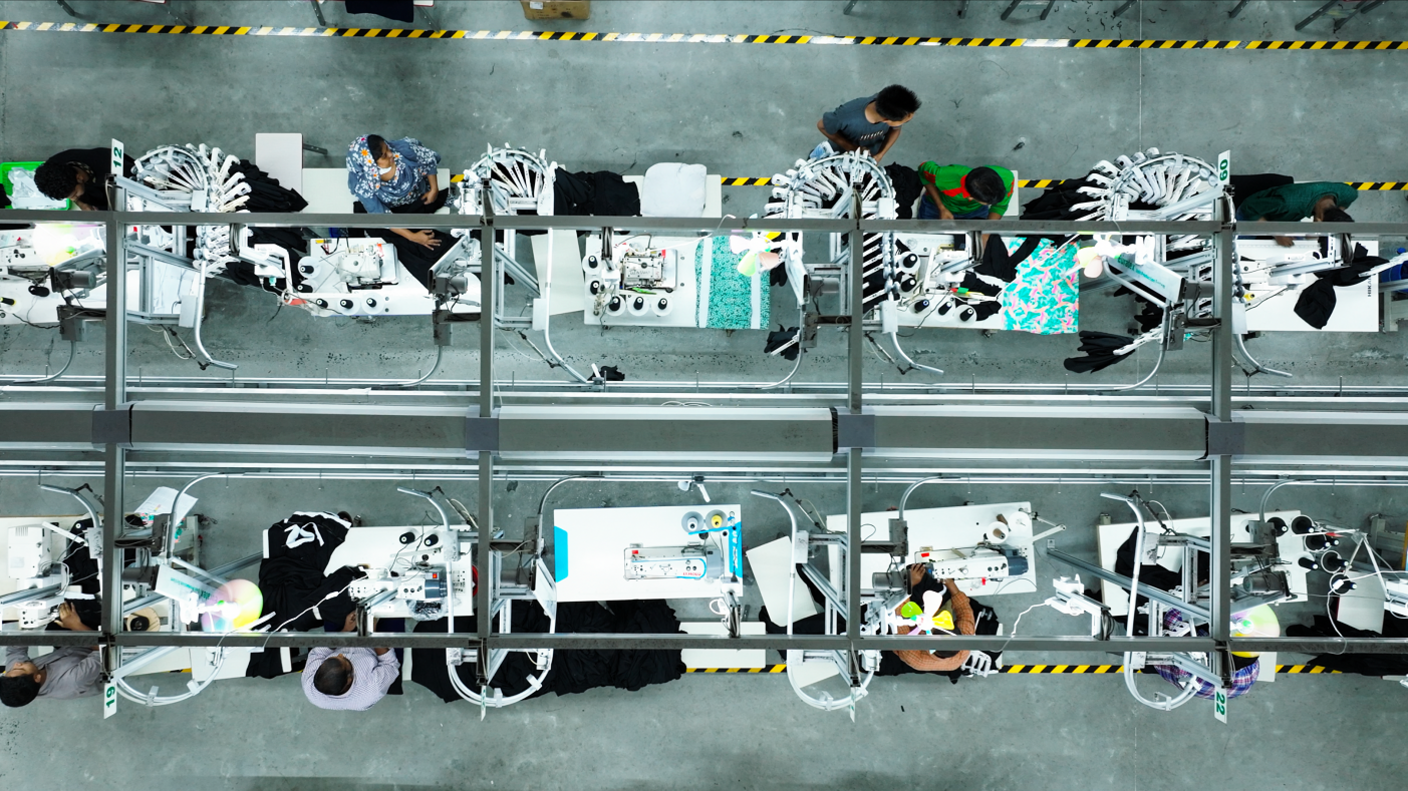 An aerial shot of a factory. The room is largely grey, with brightly-lit white work benches. People sit at each work bench. There is yellow and black warning tape on the floor.