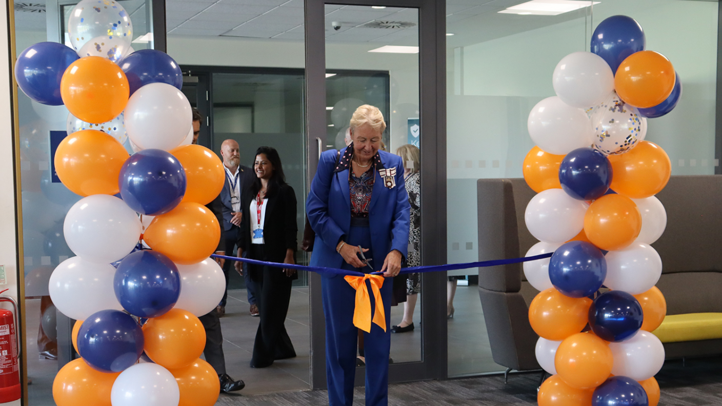 Julie Spence cutting a blue ribbon at a doorway that is surrounded by a multicoloured balloon arch.