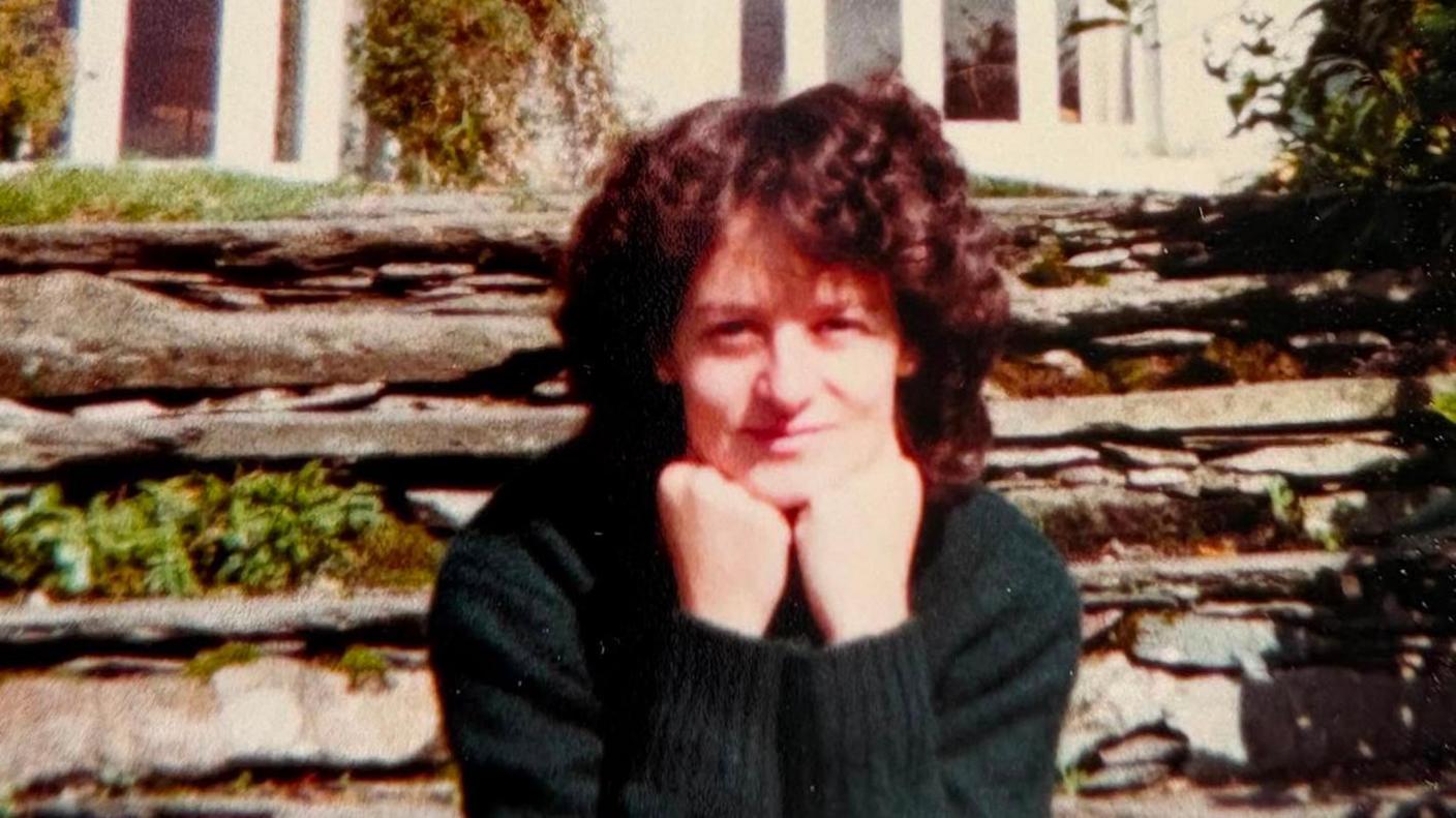 A woman sits looking at the camera with her chin resting on her balled fists. She is slightly smiling. She has curly dark brown hair and is wearing a dark green jumper. Behind her is a stone wall with some green plants growing out of it and behind that is a white house.