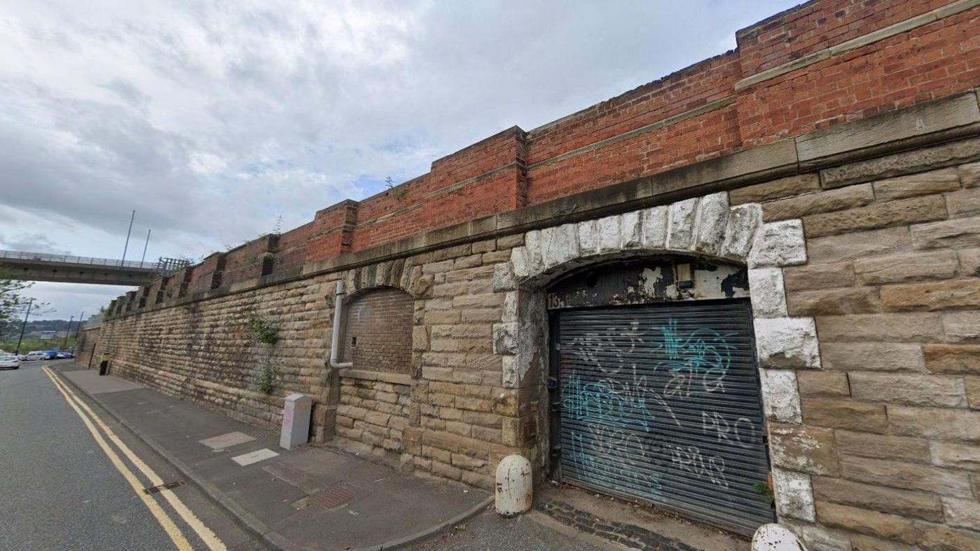 General view of Pottery Lane. There is graffiti on a garage door which is on stone wall. The Redheugh Bridge can be seen in the background.