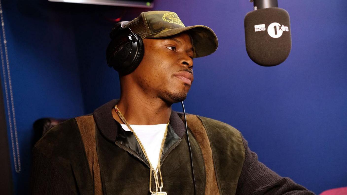 Fekky is in a Radio 1Xtra studio, with a microphone in front of him. He is wearing a camouflage cap and dark headphones. He is wearing a gold chain and dark green jacket.