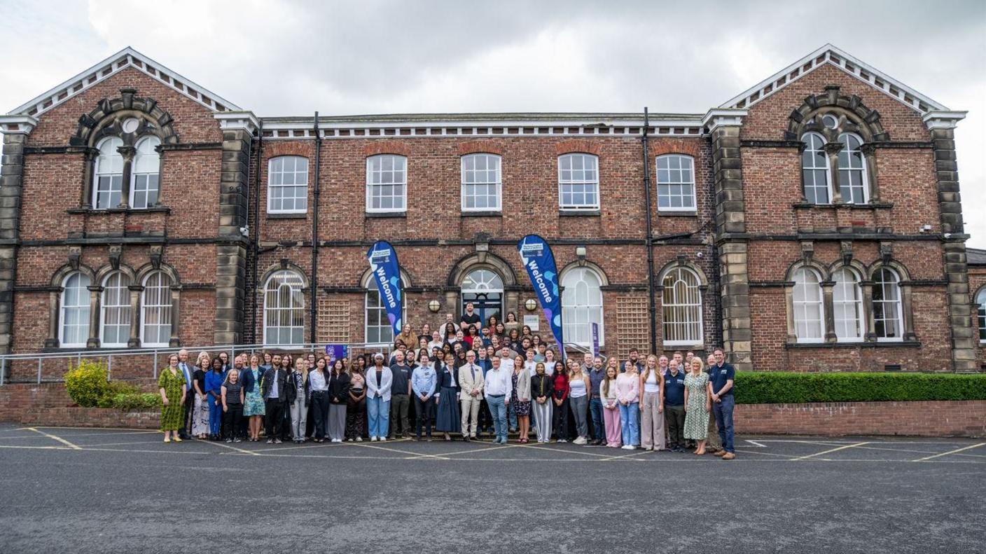 A crowd of staff and students standing outside Pears Cumbria School of Medicine. The school is based at the University of Cumbria's Fusehill Street campus in Carlisle. It is a two-storey redbrick building.