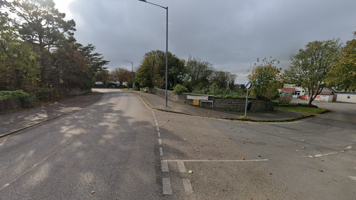 A single lane road with a junction leading off to the right. There is a brick wall running along the right side of the road and trees to the left side.