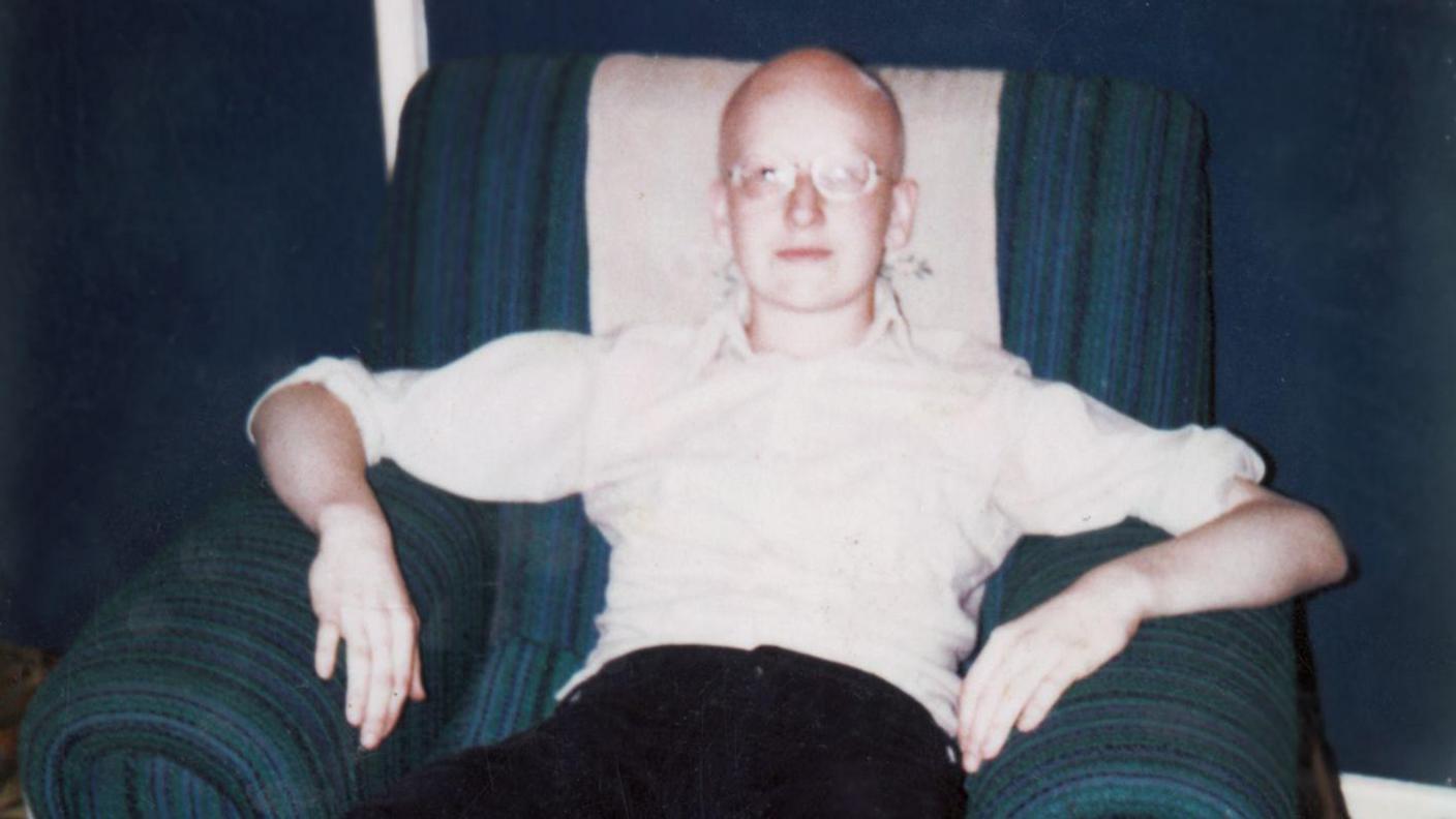 A teenage Dave Myers sits on a blue armchair. He has no hair and is wearing a white shirt, dark trousers and glasses. He looks serious and is staring at the camera.