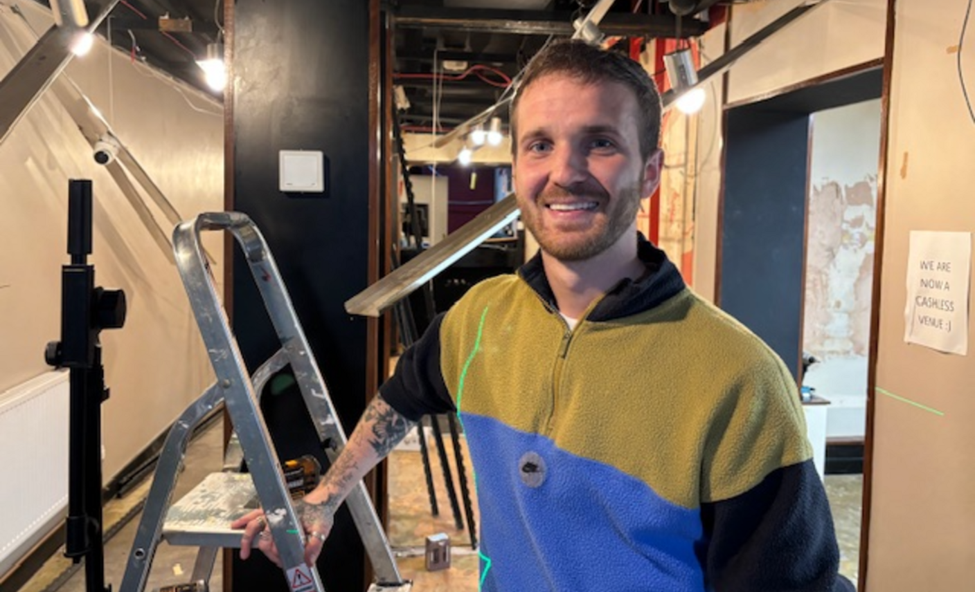 Andrew Taylor. He has short dark hair and facial hair. He is pictured next to a ladder in a corridor, looking directly at the camera and smiling.