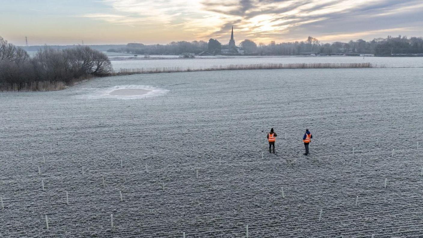 Two men in hi-vis in a field frosted with ice where trees are being planted in Lunt with a church spire in the background