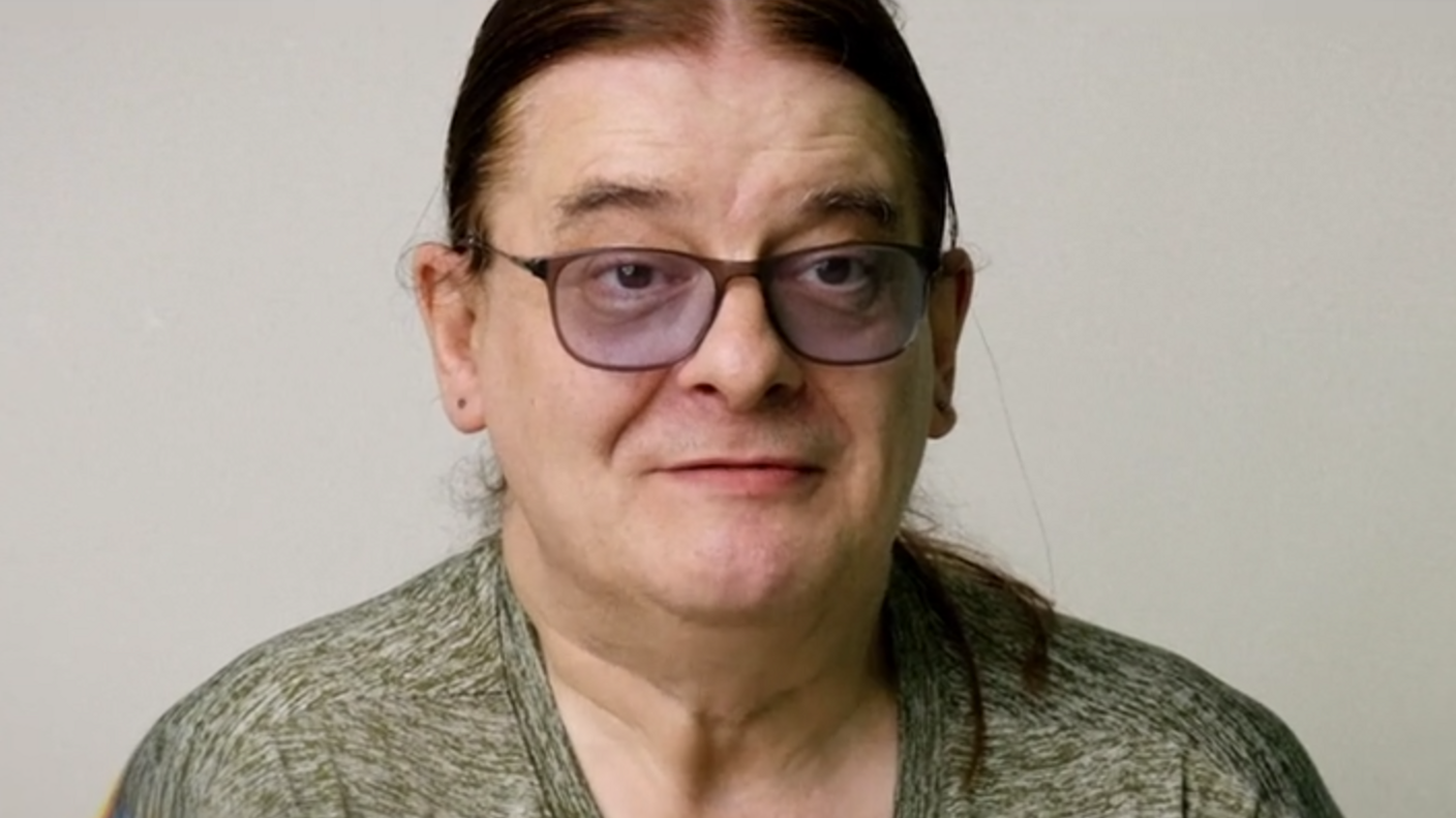 Howard, a middle-aged man with long, tied-back hair and tinted glasses. He's stood against a white background, being interviewed for the film.