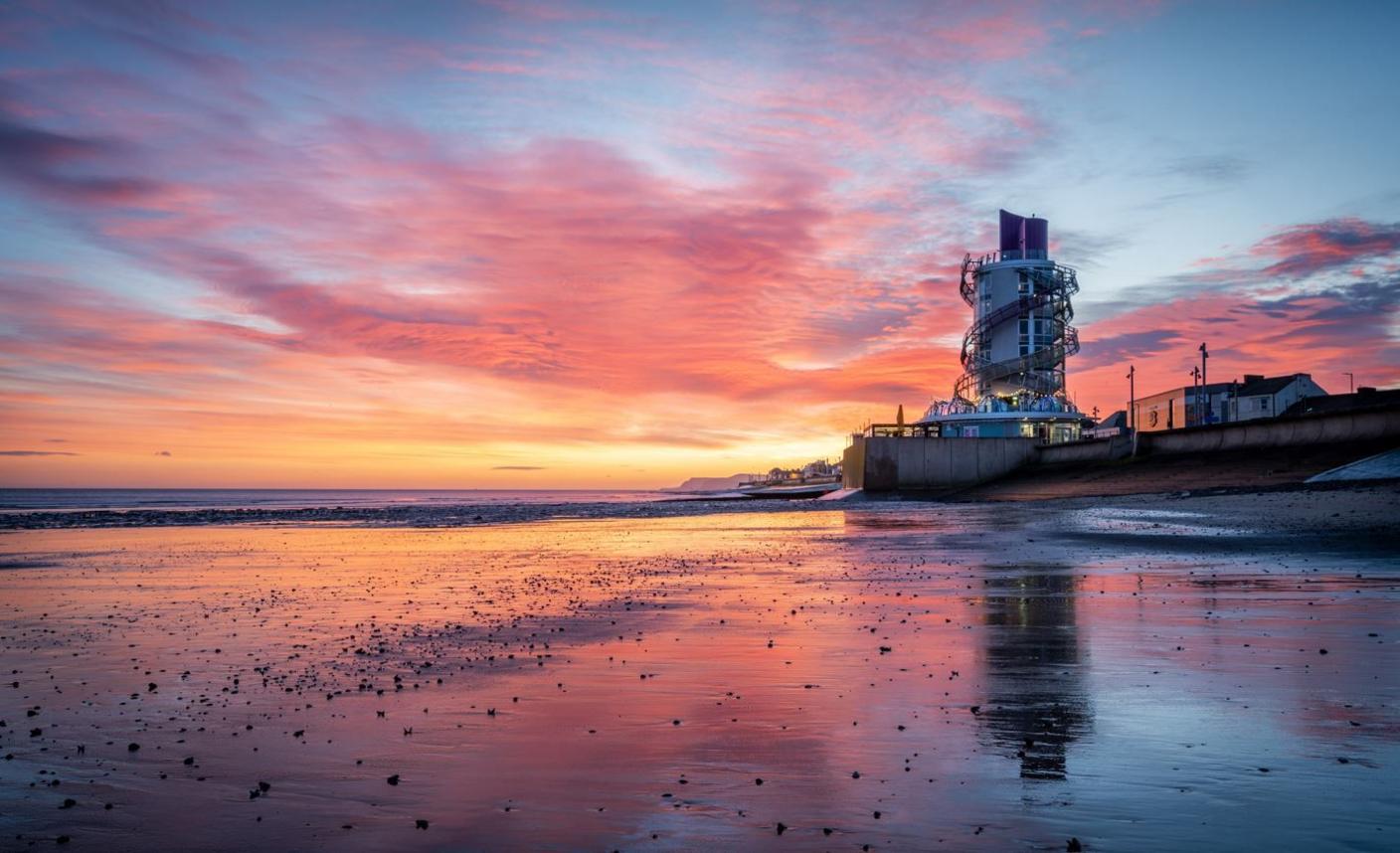 A round tower building stands next to a beach with a pink and blue sky reflected on the sand.