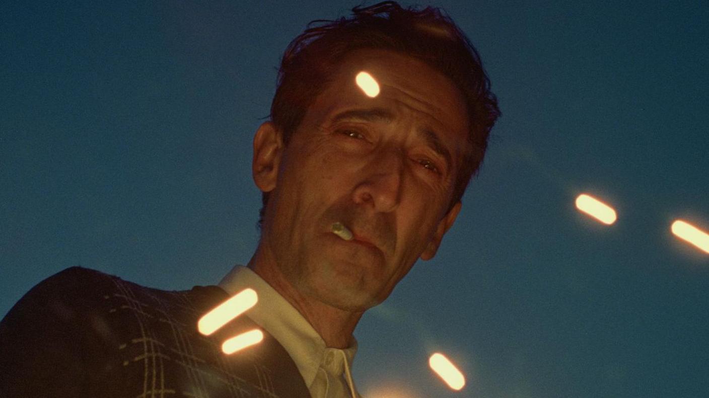 Adrien Brody as László Toth in The Brutalist. He is wearing 1950s-style clothes and is smoking a cigarette. Lights are reflecting on his face and clothing