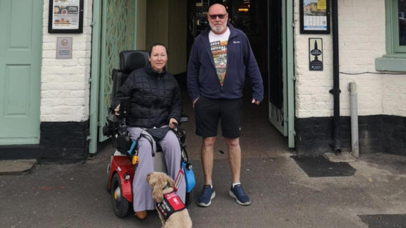 A woman in a powered wheelchair is next to a man, outside a pub. They have a dog in front of them, wearing a red and black jacket.