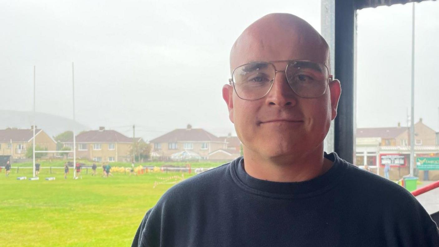 Steelworker Adam Williams, with glasses and wearing a sweatshirt, and standing near a rugby field looks into the camera