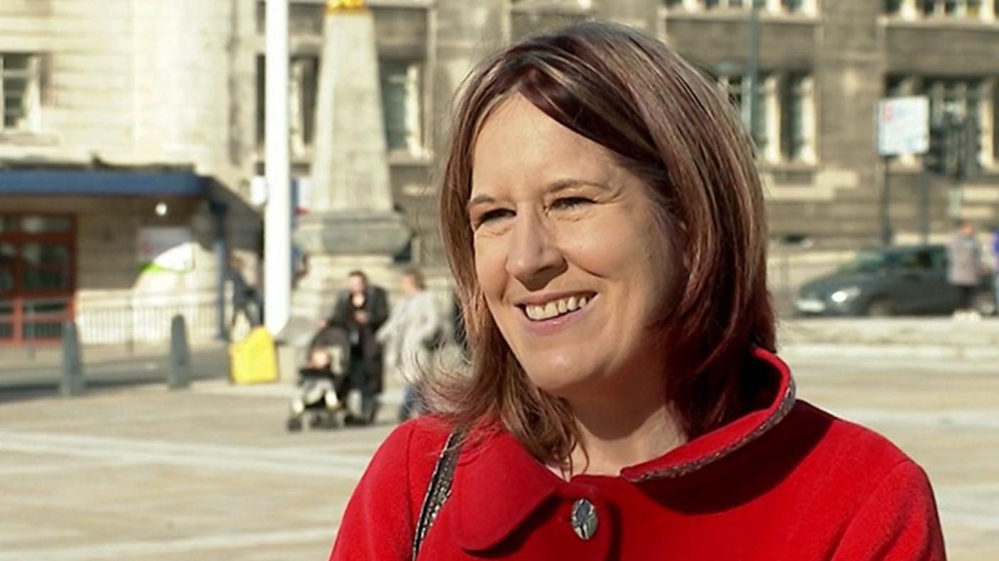 Ms Venner wears a red coat and has a handbag slung over her right shoulder. She has mid-length brown hair and is smiling at the camera. Two people pushing a pram and two buildings can be seen in the background.