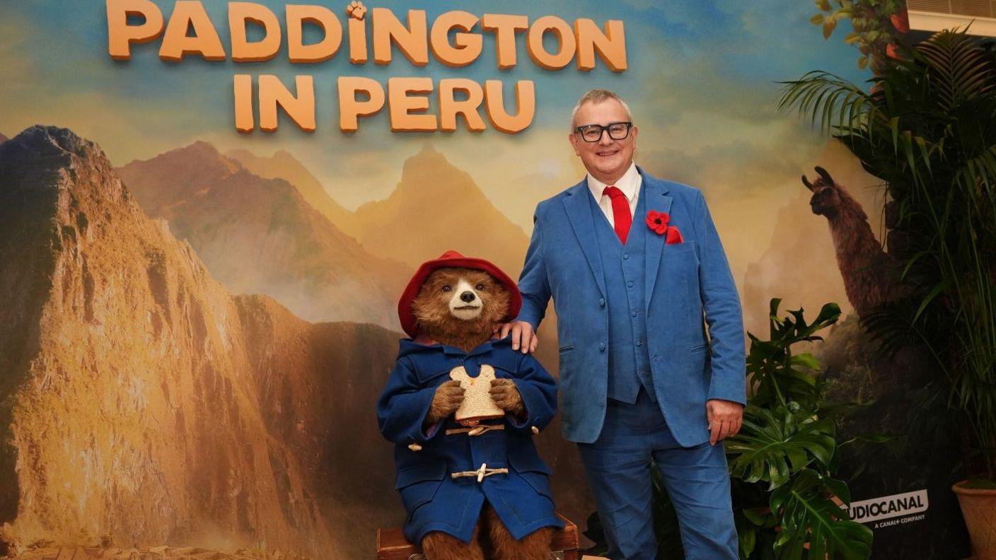 Hugh Bonneville poses for a photo with Paddington Bear in front of a large poster advertising the new film Paddington in Peru. He wears a light blue suit with matching waistcoat and trousers, a bright red tie and a red poppy on the lapel. He has thick dark glasses and is smiling. paddington is holding a sandwich with a chunk bitten out, and wears his blue duffel coat and red hat. The poster shows mountains and jungle in the background.