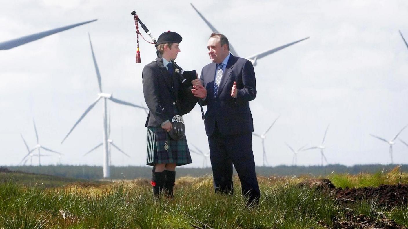Former first minister of Scotland, Alex Salmond, is talking to a bagpiper. They are standing on grass in front of a large wind farm.