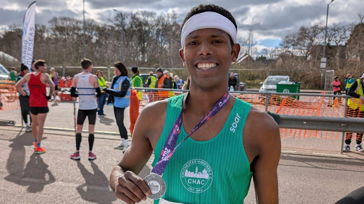 Mohammed Elbayan wearing a green vest and holding a large medal. He has a white headband on and is standing at the finish line of a run, where several runners and staff are milling about.