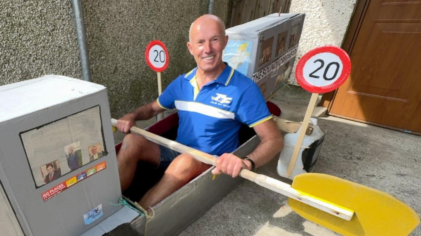 Dave Kelly, wearing a blue T-shirt, sits in a metal bathtub which is on grey concrete. He holds a paddle and smiles. Two grey cardboard boxes have been attached to the front and back. Two paper plates have been turned into 20mph speed limit signs on either side of him.
