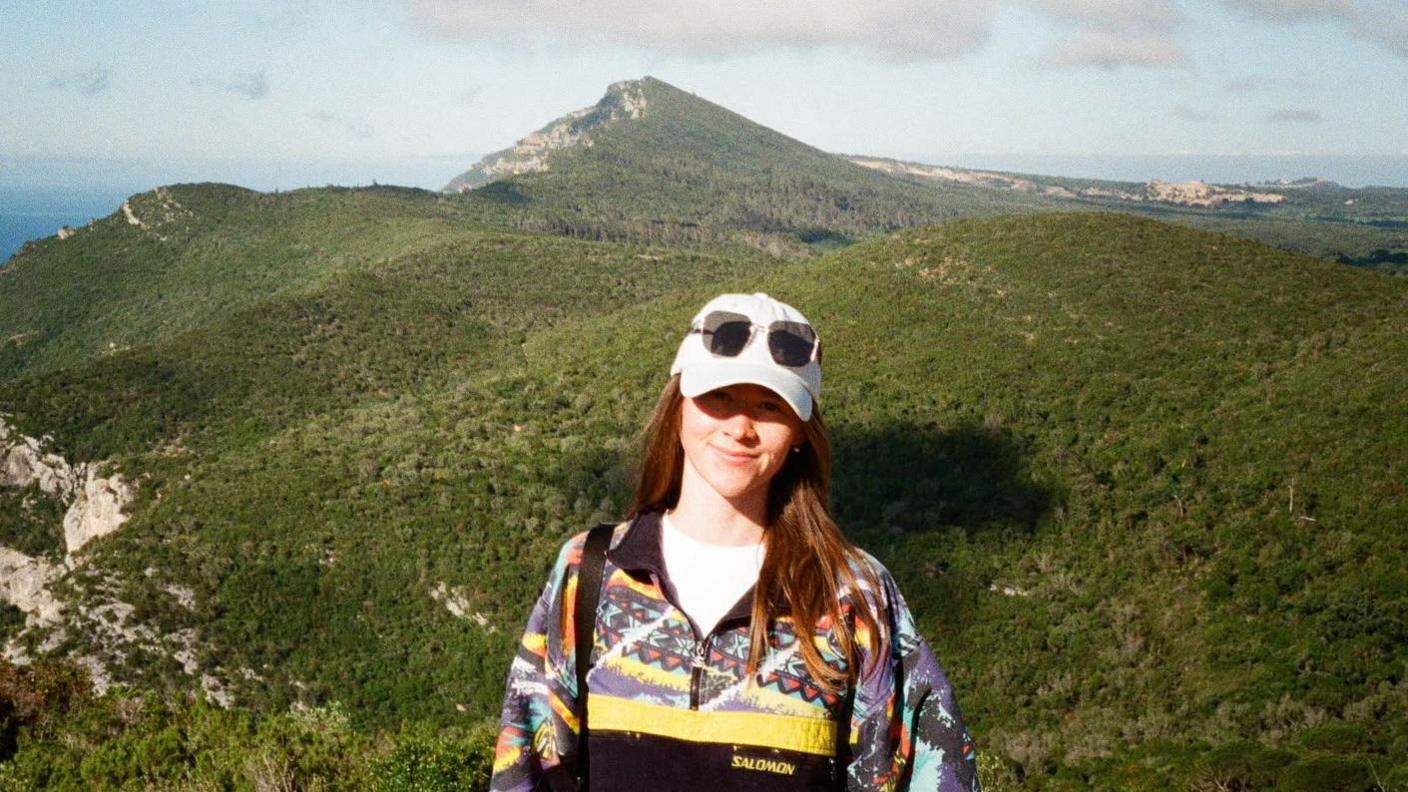 Shannon yn sefyll ar ben mynydd. Mynyddoedd gwyrdd tu ol iddi