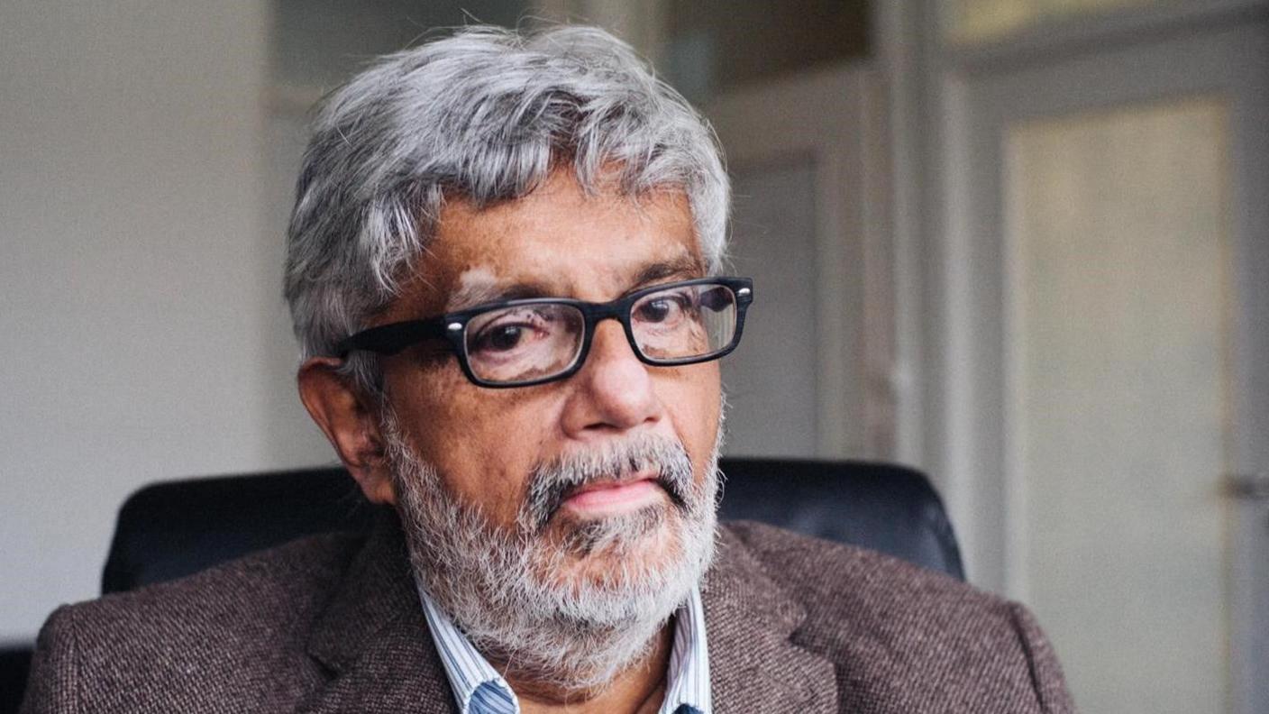 Viraj Mendis is pictured in 2019 ahead of his last interview. He has grey hair and a beard and is wearing glasses. He is wearing a blazer and striped shirt. 