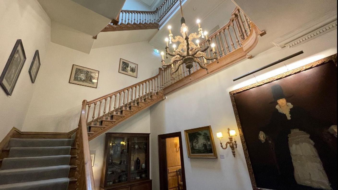 There is a large wooden sweeping staircase with a hanging chandelier. There is a large portrait of Dorothy Elmhirst dressed as a pilgrim, hanging on a wall.