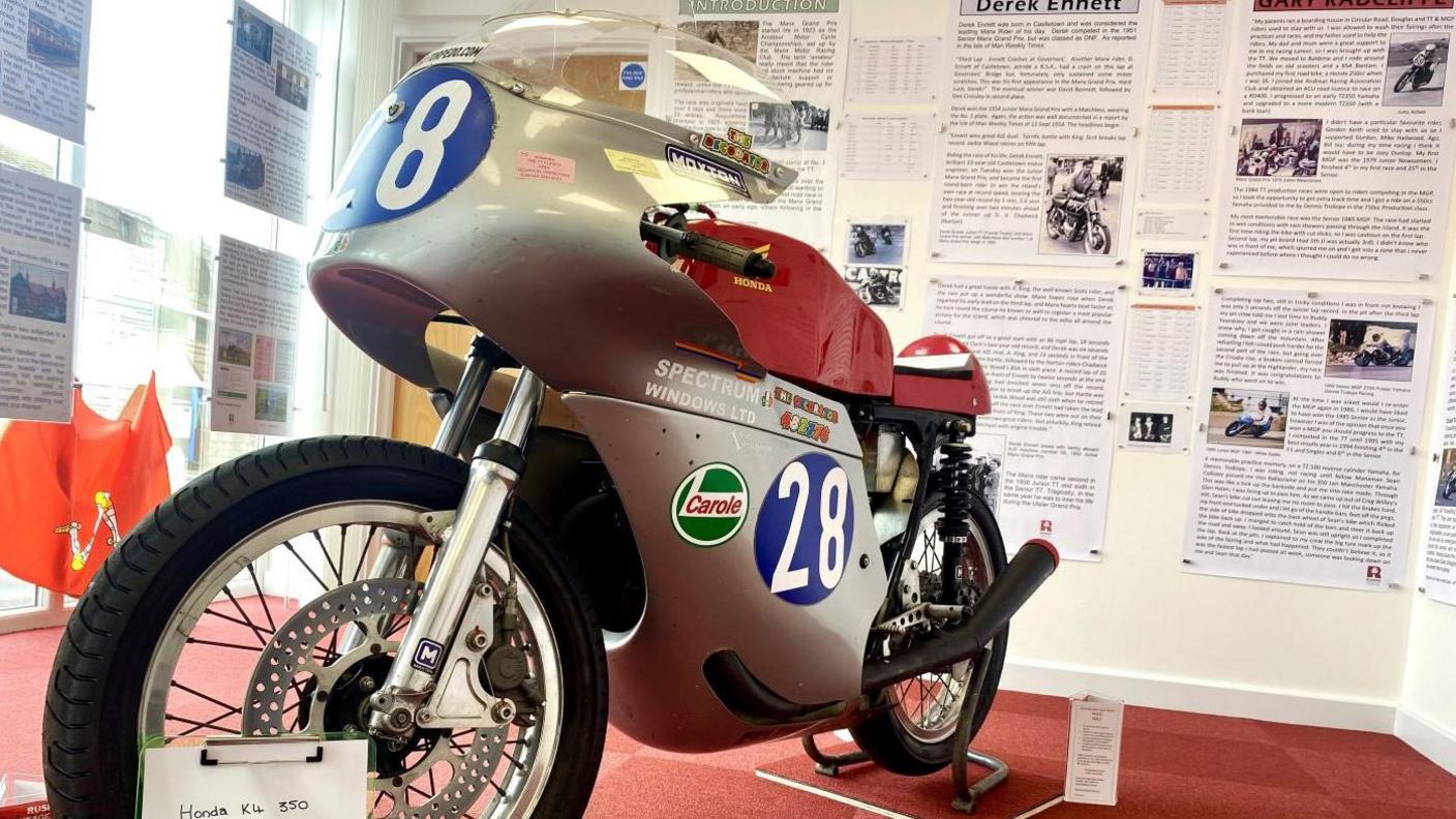 A classic silver Honda K4 350 racing bike with a red tank and saddle, which features the number 28 in white painted in a blue circle on the side and front. The motorbike in on a stand on a red carpet with display boards featuring information about riders on the wall behind.