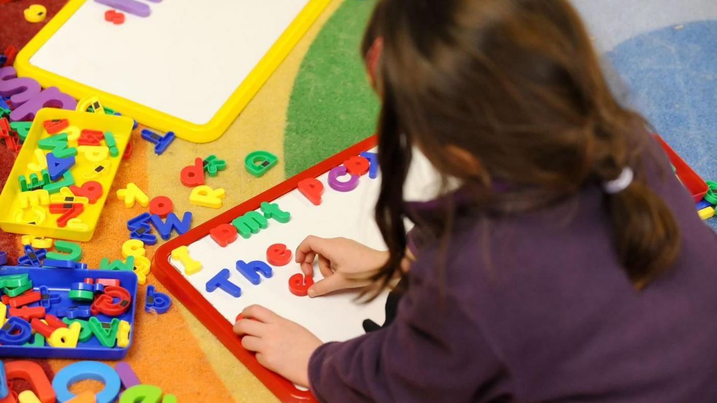 A stock image of a young child who is unidentifiable playing with some magnetic letters on a whiteboard. 