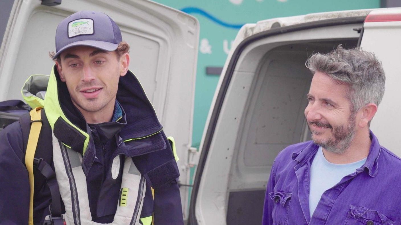 Martin Marescaux standing next to Steve Oxford who is smiling at Martin. Martin is wearing a dark lifejacket and cap. Steve has a full beard and is wearing a blue shirt.