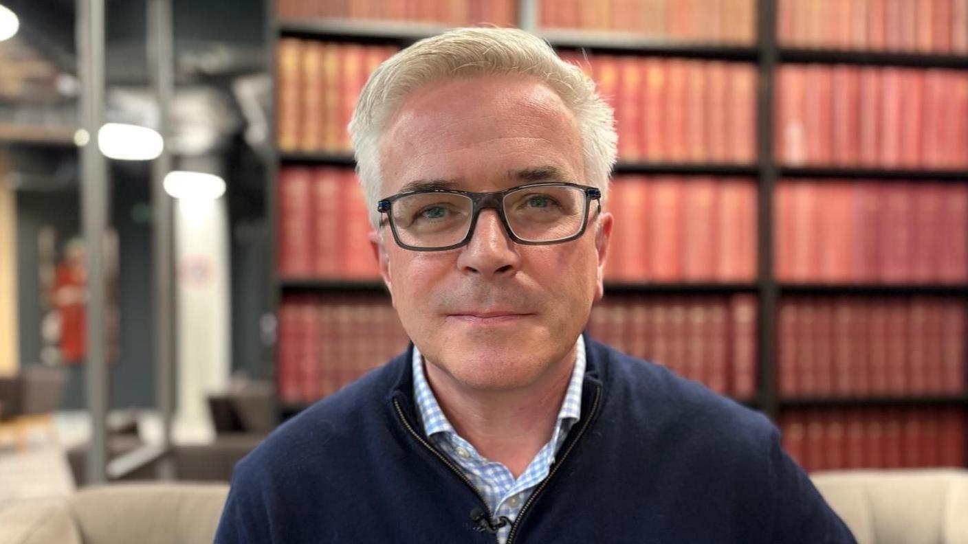 Alun is wearing dark-framed glasses and has a neutral expression. He is wearing a light blue checked shirt with a navy sweater. Behind him, blurred, are red books on a large bookshelf. It is a head and shoulders shot of him.