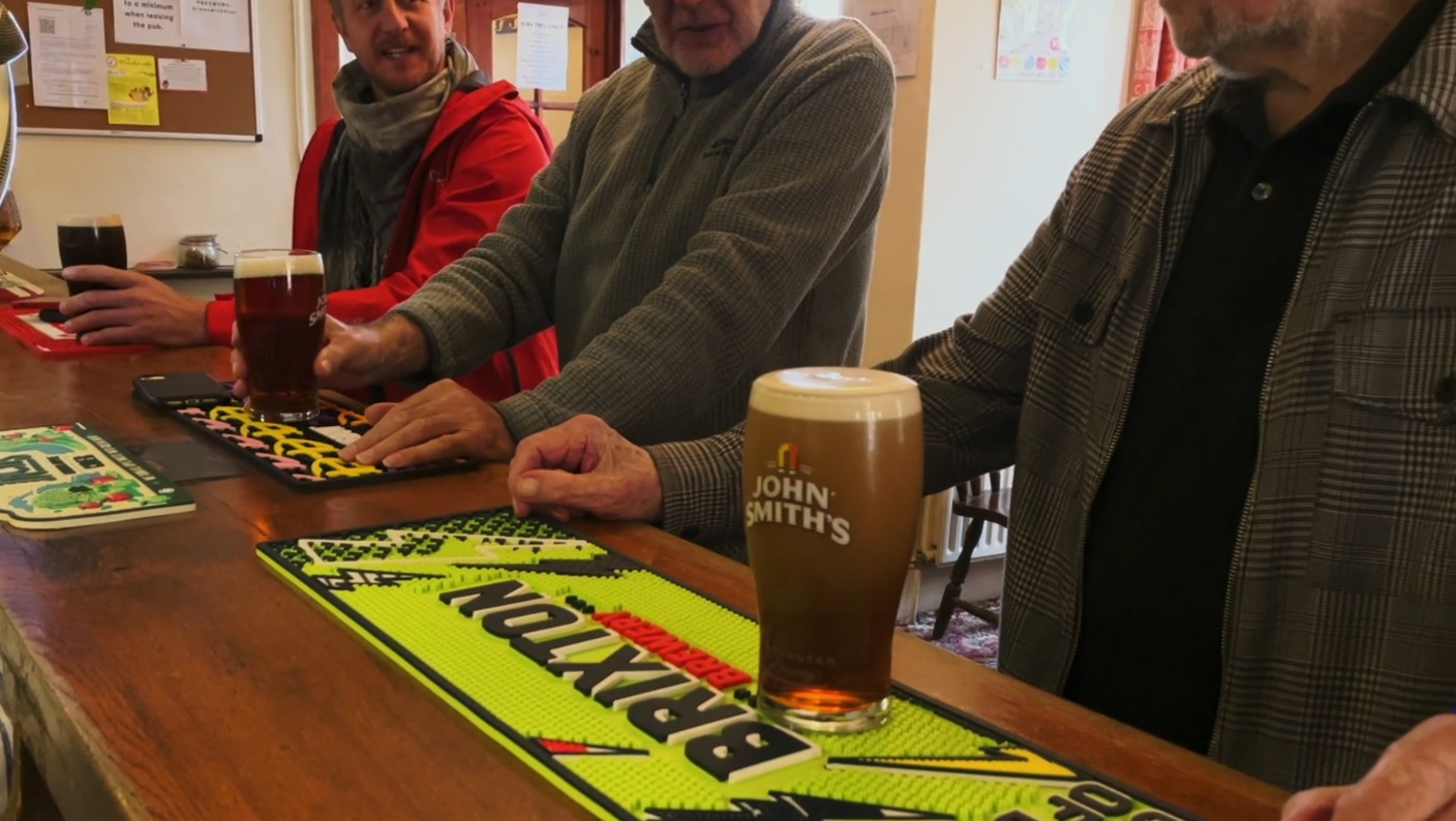 The image shows men at a pub bar with pints of beer, including a "John Smith's" pint in the foreground. 