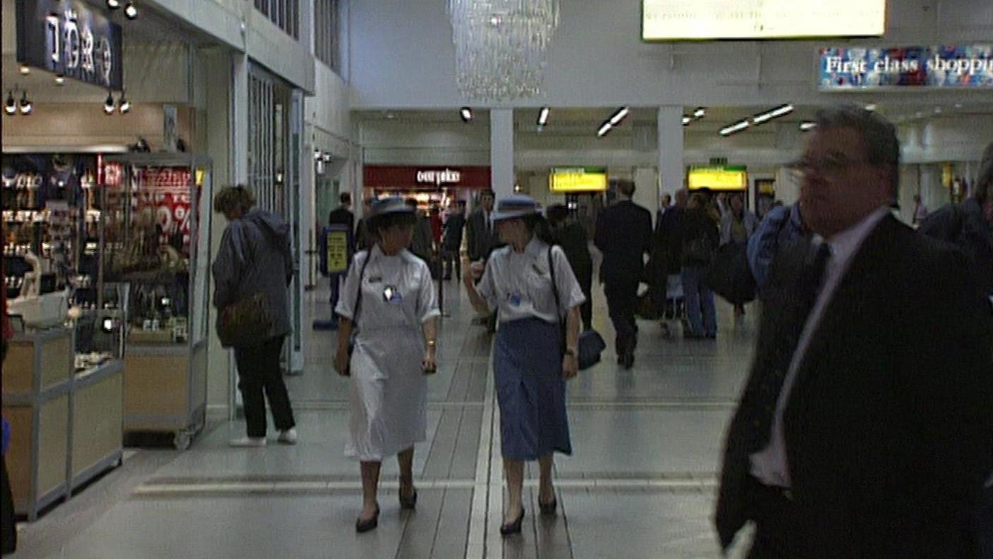 Two women walk through Terminal 1, they were white and blue uniforms, the pass under a large chandelier 