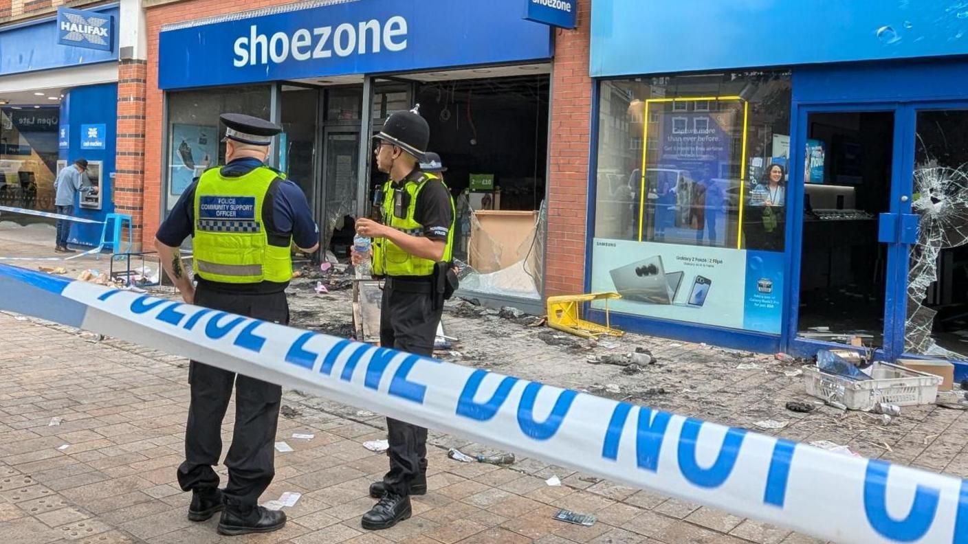 Two police officers stand between a blue-and-white cordon and a badly damaged Shoezone shop in Hull city centre. Windows are broken and there is charred debris on the ground.