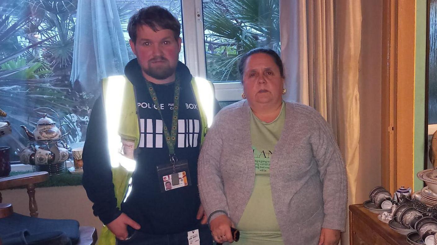 Maria Cooper and her son Paul stand in a living room. Maria has tied-back, brown hair and wears a green top and grey cardigan. Paul has brown hair and a beard and wears a high-vis jacket and a dark top with the words "police box".