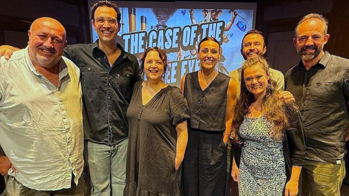 Steve Spiers stands left and is grinning and wearing a light blue linen shirt, next to his son Lewes Roberts, who has his arm around him and is wearing glasses, navy shirt and blue jeans. Ruth Jones is next to him and is wearing a black linen dress and has her hair tied back. Suranne Jones is next to her, with her hair combed back and in a black linen trouser suit; Rachael Cummins in a blue print dress and cardigan stands in front of Ryan Starling, whose hair is combed back, and to the side of Laurence Akers, who is also smiling and wearing a black shirt and khaki trousers. Behind them is a background that says ' The Case of...'