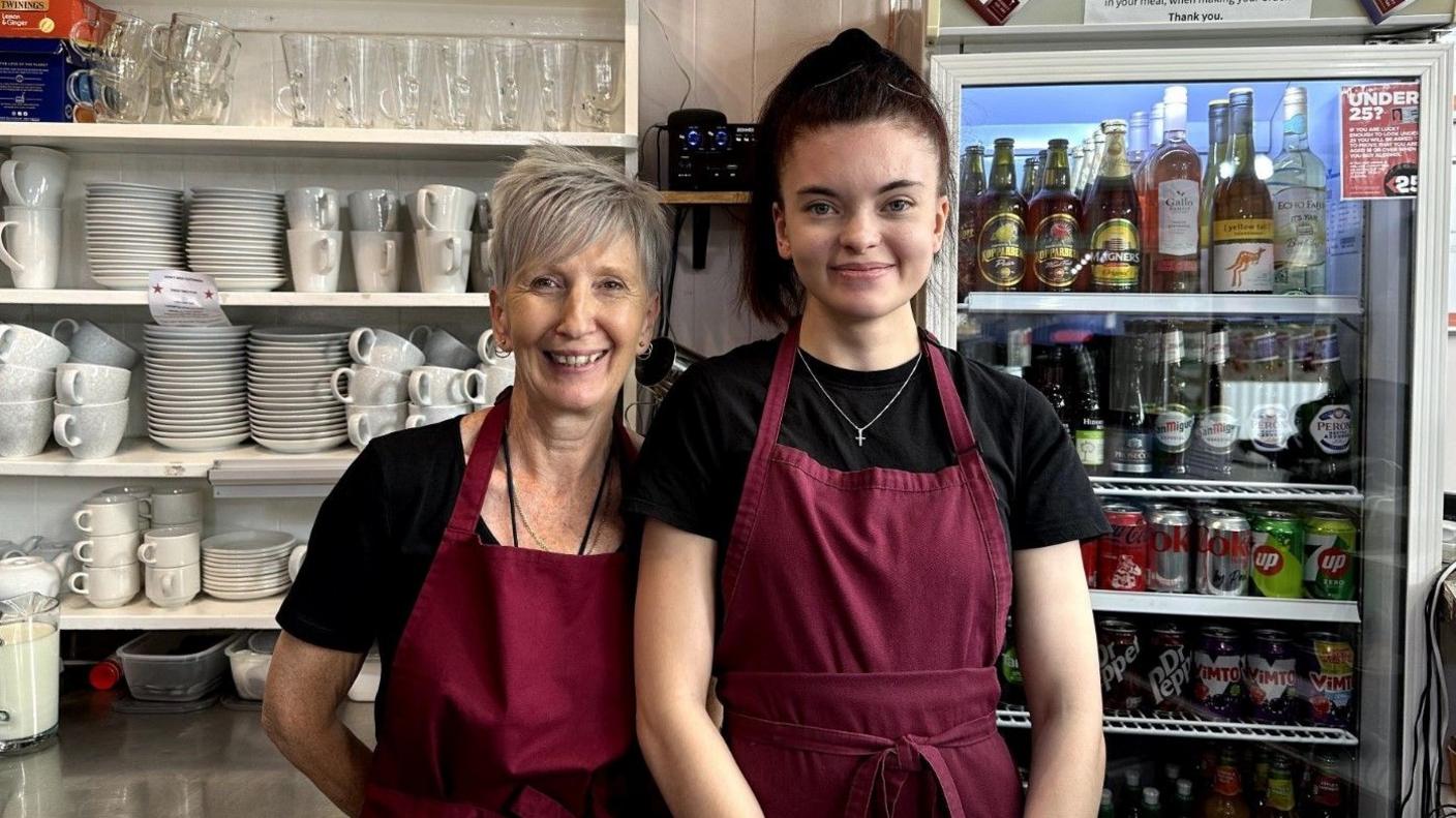 Two women wearing maroon aprons standing behind a cafe counter, smiling