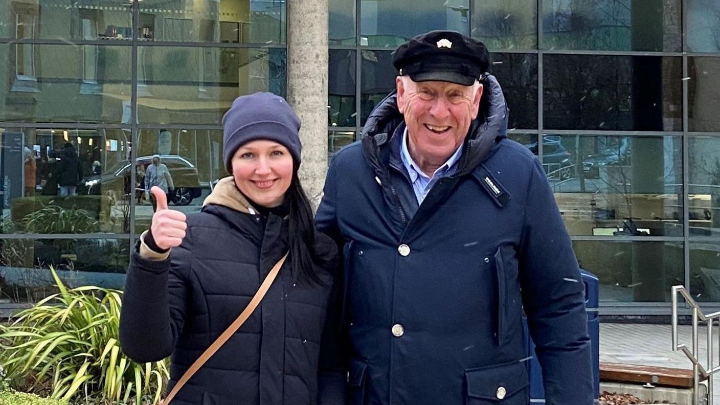 An older man and a younger woman, both wearing navy blue coats, stand with one arm around each other. The woman is giving the photographer a thumbs-up.