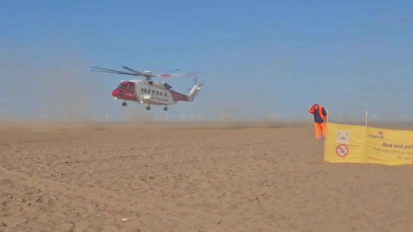 Sand is whipped up as a large red and white helicopter comes into land on a sandy beach under a blue sky. To the right, a man wearing orange hi-vis overalls bows his head as he stands next to a yellow Lifeguards sign.