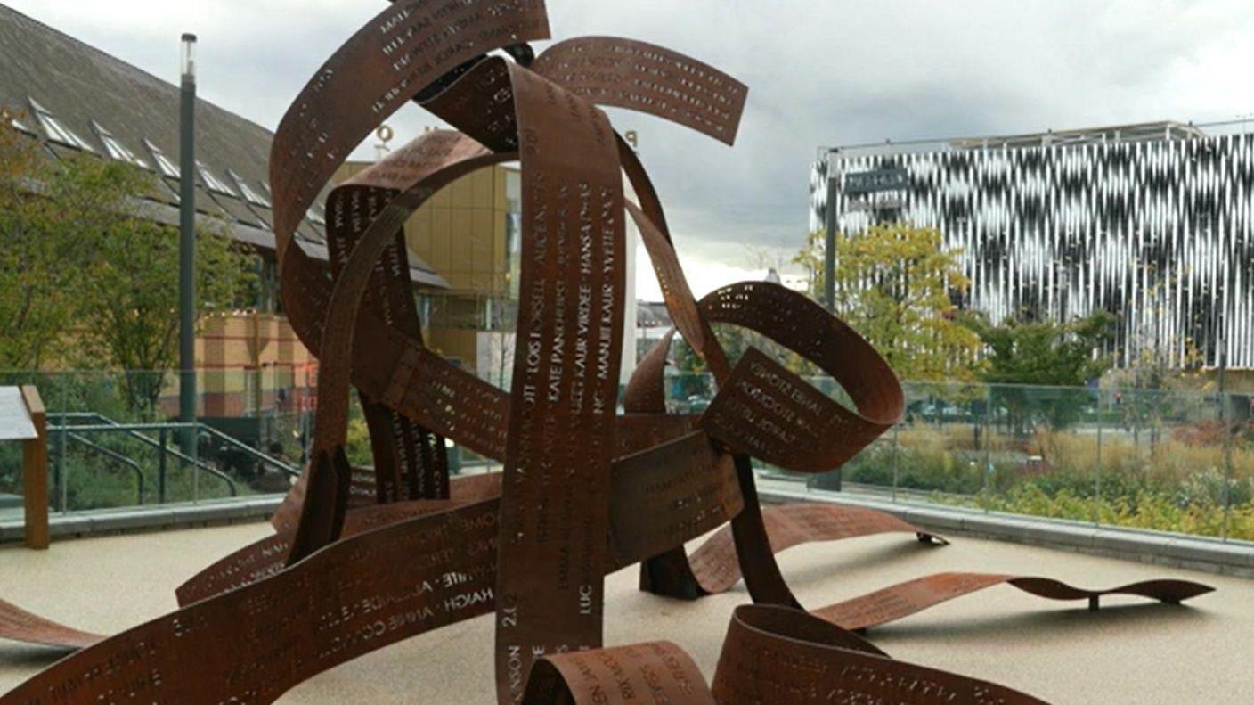 The corten steel artwork rises up from the ground. The five ribbons carry the names of the hundreds of women and are entangled as they meet at the top.