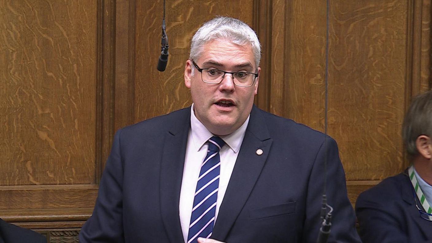 Gavin Robinson speaking in the House of Commons. He has white hair, wearing glasses, a navy blazer, white shirt and blue and white stripped tie, a gold pin is on his lapel. Wood panelling is behind him.