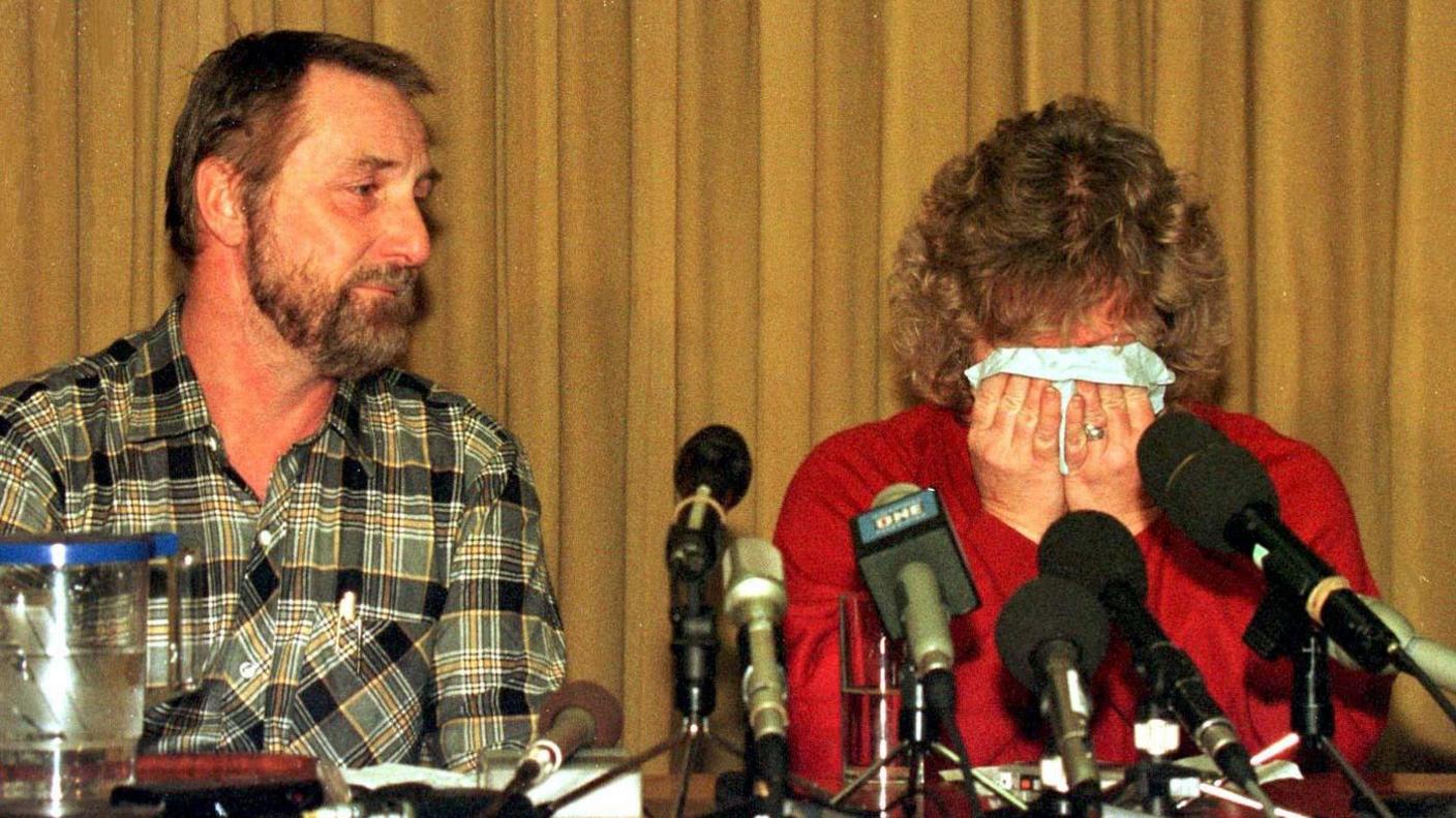 Paul and Janet Betts sitting in front of microphones lined up on a desk at a press conference. Janet is covering her face with a tissue while crying, head in hands. Paul is also visibly emotional while looking at his wife.