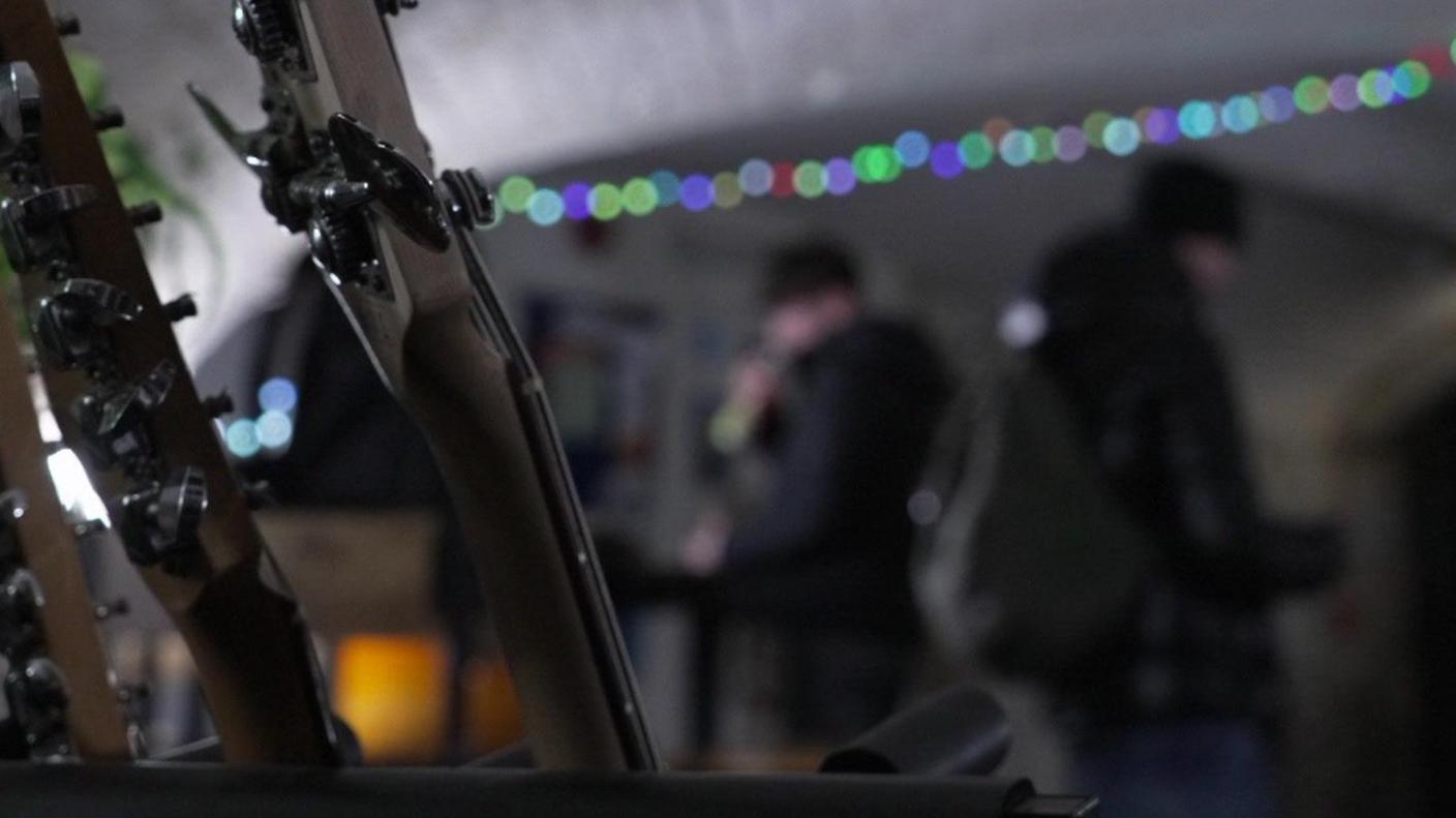 Three guitar necks are in the foreground. The background is very blurred, but two young men wearing black beanie hats can be made out. One of them is holding a microphone to his mouth. You cannot make out their faces. There is a string of multi-coloured fairy lights across the ceiling.