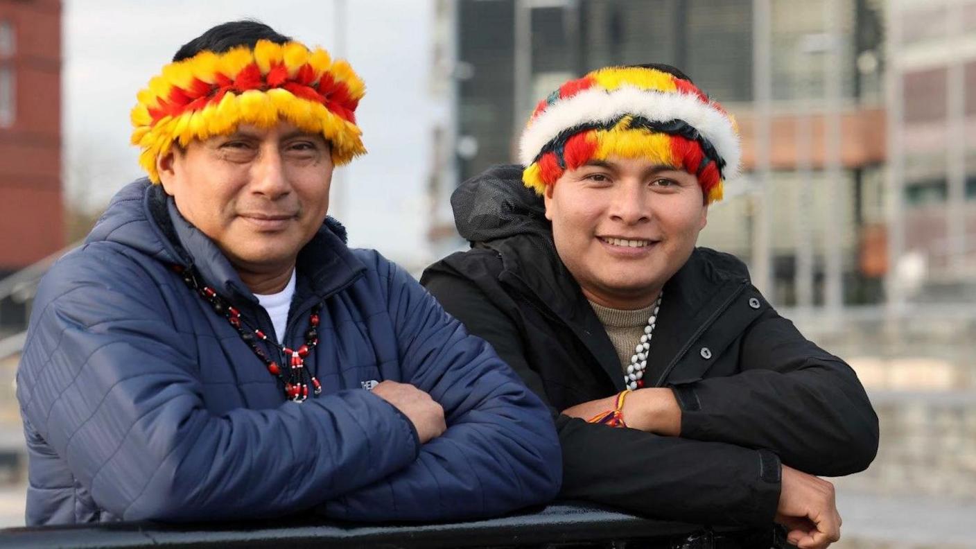 Teófilo Kukush Pati, President of the WampÃs Nation - an indigenous nation from the Peruvian Amazon – and Tsanim Wajai Asamat standing outside the Senedd in Cardiff Bay. The man on the left has his arms crossed and wears a navy puffer coat. He has a black and red beaded chain. And a yellow and red feathered headdress. He looks at the camera. The man next to him also has his arms crossed, and has a black jacket on. He smiles at the camera and has a yellow, red and white headdress.